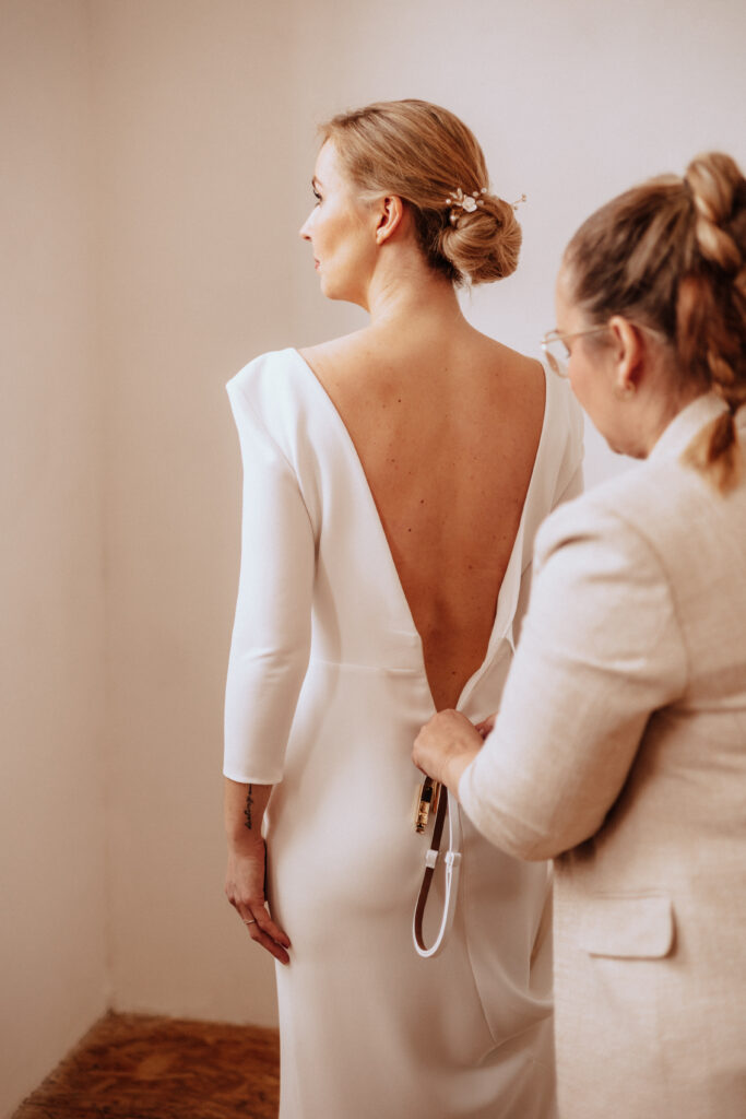 In einem sanft beleuchteten Raum in Brandenburg schließt eine Frau das weiße, hinten offene Brautkleid mit einem Reißverschluss, umgeben von eleganter Hochzeitsdeko.