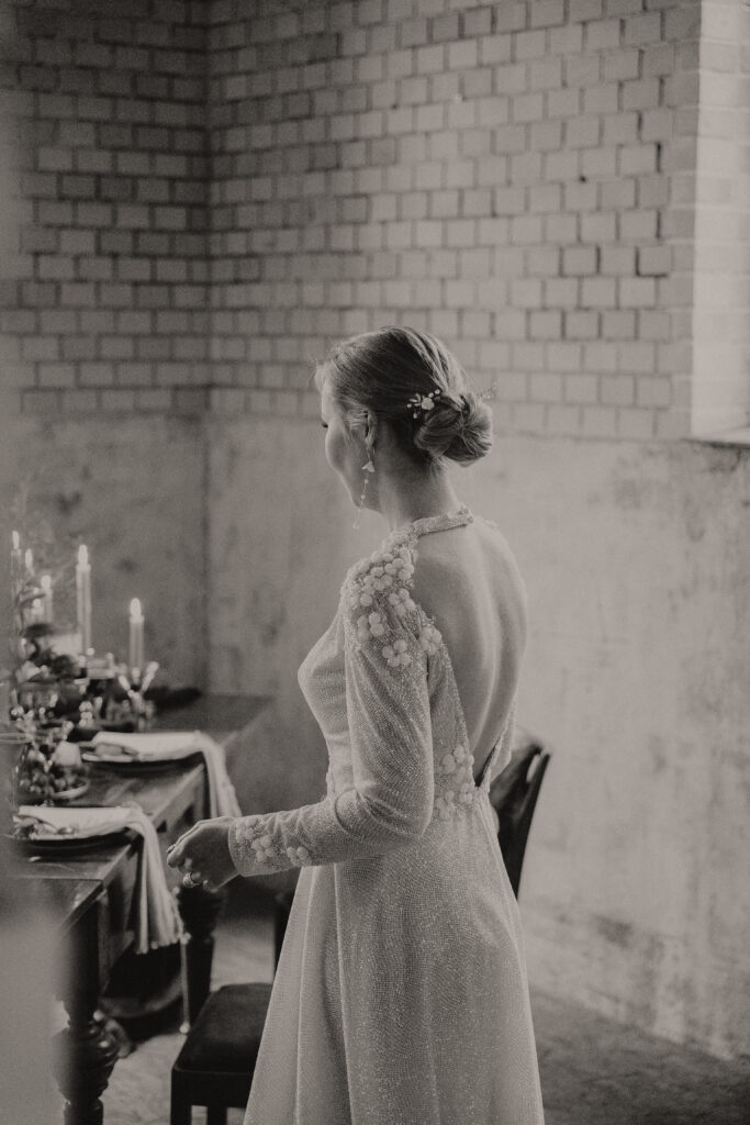 Eine Braut in einem langärmeligen Kleid mit offenem Rücken steht an einem mit Kerzen beleuchteten Tisch in Berlin und schaut in die Ferne - eine elegante Szene, die von einem talentierten Hochzeitsplaner kuratiert wurde.