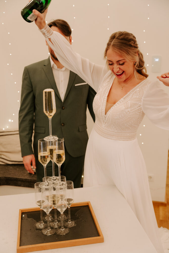 Eine freudige Braut schenkt Champagner in einen Turm von Gläsern ein. Ihr Bräutigam steht neben ihr, umgeben von einer eleganten Hochzeitsdeko auf ihrer Berliner Feier.