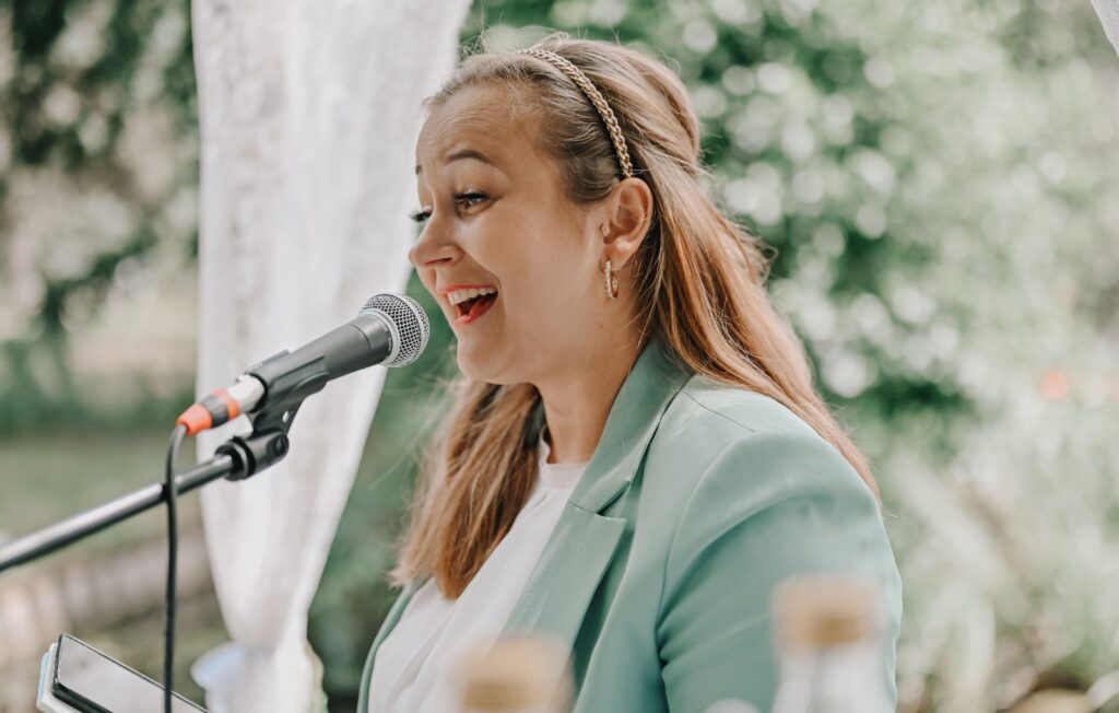 Strahlend in einem grünen Blazer spricht oder singt eine Freie Traurednerin in Berlin in ein Mikrofon im Freien und lächelt dabei fröhlich.