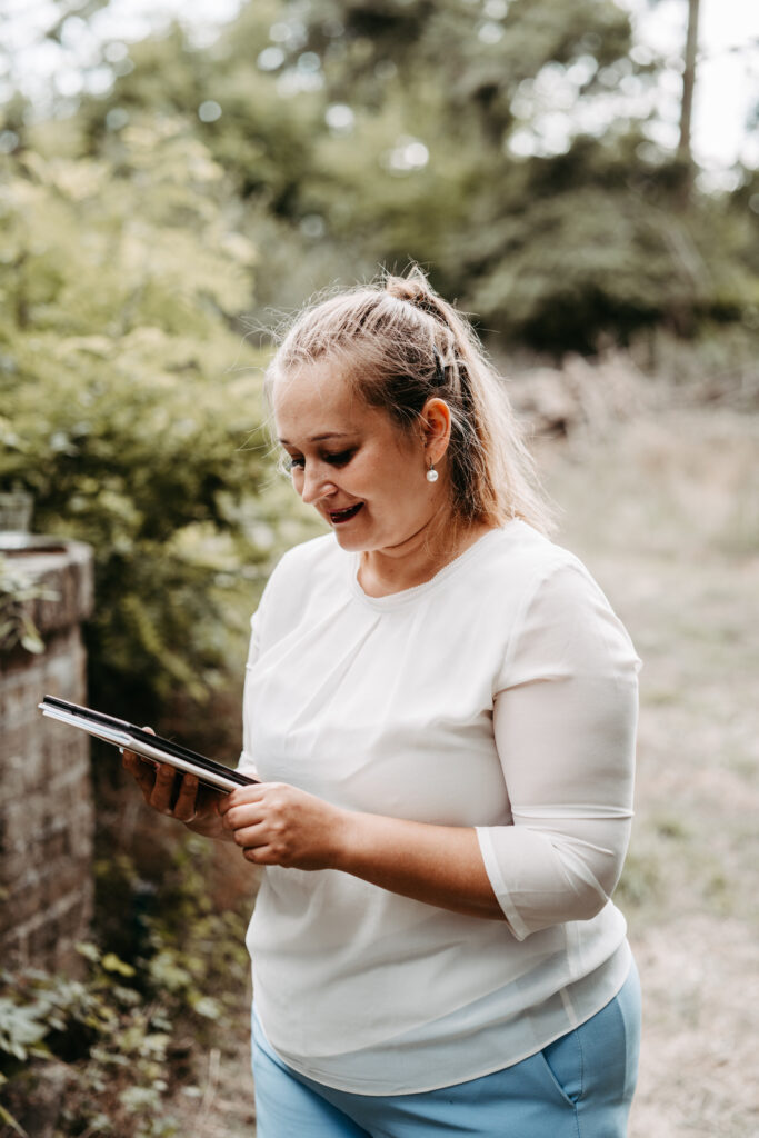Eine Frau lächelt, während sie im Freien in Brandenburg ein Tablet benutzt, umgeben von grünem Laub und natürlichem Licht.