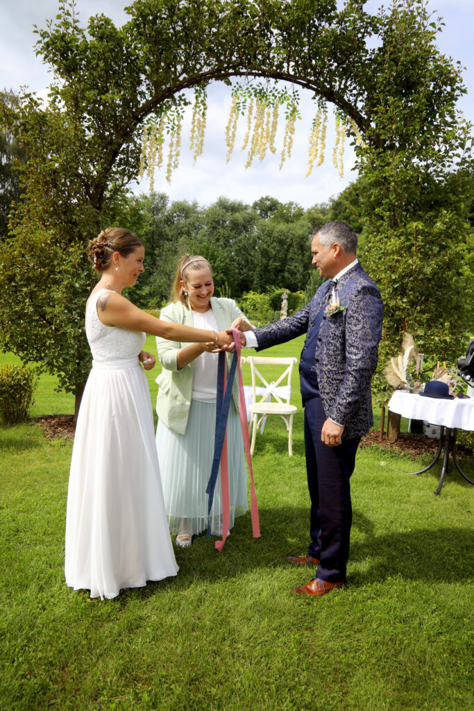 Braut und Bräutigam stehen unter einem blumengeschmückten Hochzeitsdeko-Bogen und halten bunte Bänder mit einem Trauzeugen auf einer grünen Wiese in Brandenburg.