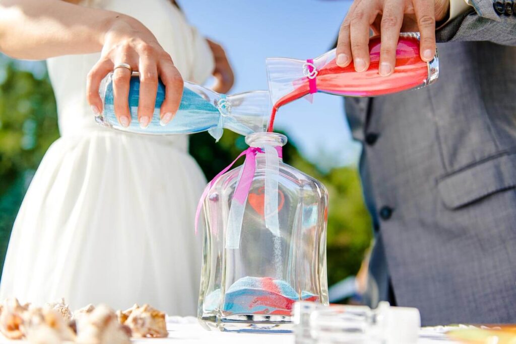 Ein Paar schüttet während der Hochzeitszeremonie blauen und rosa Sand in ein Glasgefäß und schafft damit einen einzigartigen Hochzeitsdeko-Moment. Dieses berührende Ritual wird oft von einem Hochzeitsplaner oder einer Freien Traurednerin empfohlen.