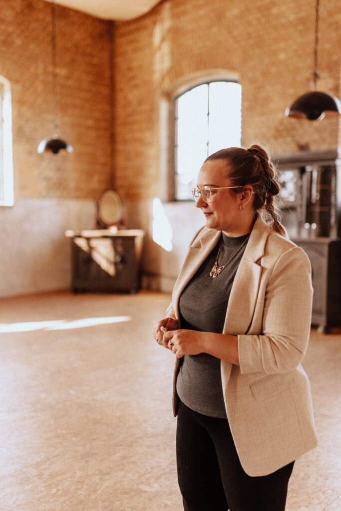 Eine Frau im beigen Blazer steht lächelnd in einem sonnendurchfluteten, großzügigen Raum mit Backsteinwänden und großen Fenstern - perfekt für eine Freie Traurednerin in Berlin, die Inspiration für eine Hochzeitsdeko sucht.