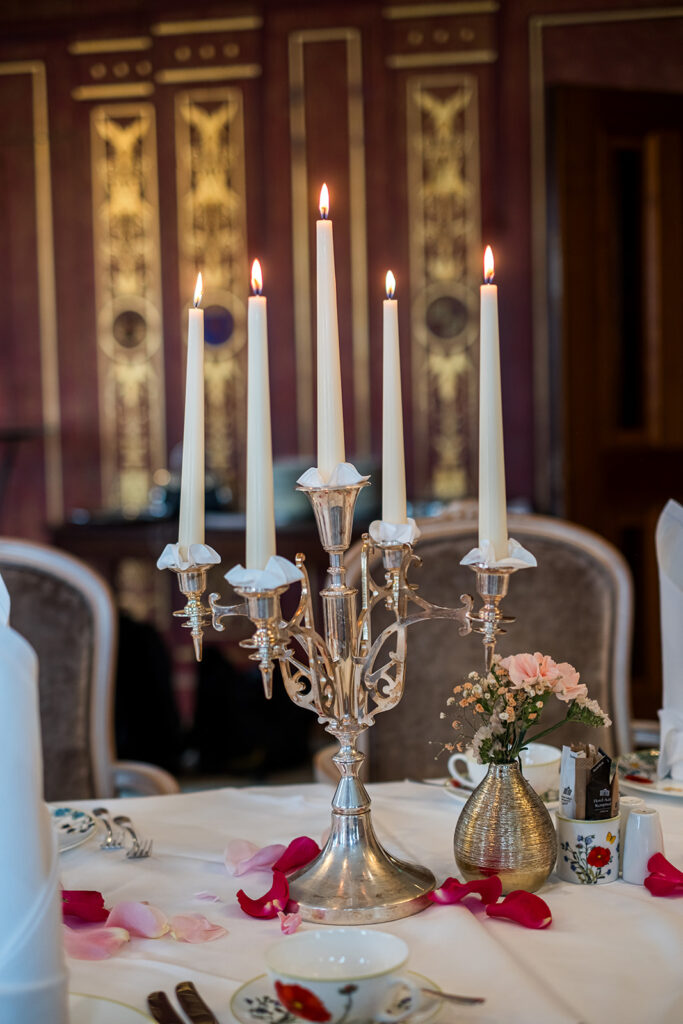 Ein silberner Leuchter mit fünf brennenden Kerzen ziert einen dekorierten Tisch im Hotel Adlon Berlin, der mit Rosenblättern und einer kleinen Blumenvase geschmückt ist.