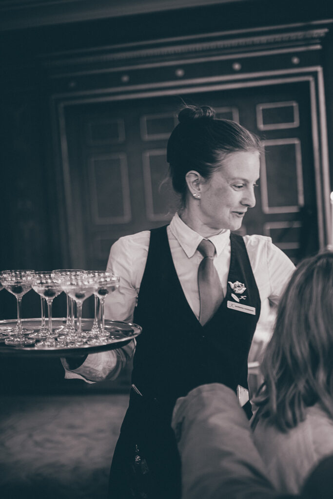 Ein Kellner in Uniform serviert einem sitzenden Gast im eleganten Ambiente des Hotel Adlon Berlin ein Tablett mit leeren Weingläsern.