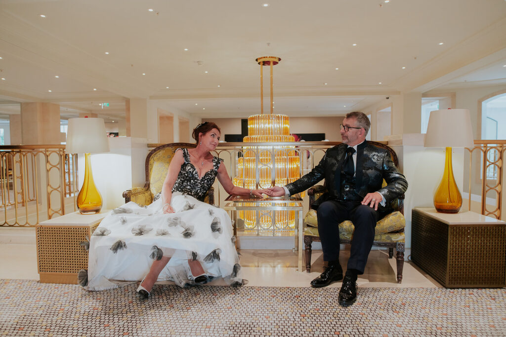 Ein formell gekleidetes Paar sitzt händchenhaltend und lächelnd auf verzierten Stühlen in der eleganten, gut beleuchteten Lobby des Hotel Adlon Berlin.