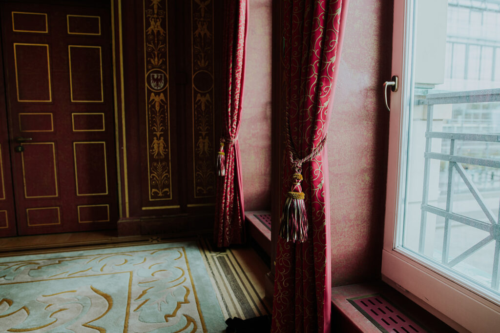 Opulente rot-goldene Vorhänge an einem großen Fenster in einem reich verzierten, klassizistischen Zimmer, das an die luxuriöse Einrichtung des Hotel Adlon Berlin erinnert.