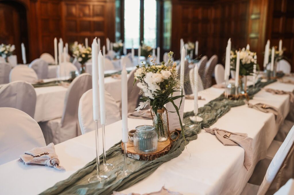 Elegante Banketttafel mit weißer Bettwäsche, floralen Tafelaufsätzen und hohen weißen Kerzen in einem holzgetäfelten Raum - perfekt für Ihre nächste Veranstaltung in Berlin mit unserem exklusiven Dekorationsservice und Dekoverleih.