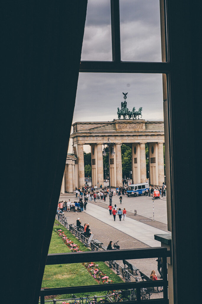 Blick auf das Brandenburger Tor und eine Menschenmenge draußen, gesehen durch ein Fenster mit einem dunklen Vorhang im Hotel Adlon Berlin.