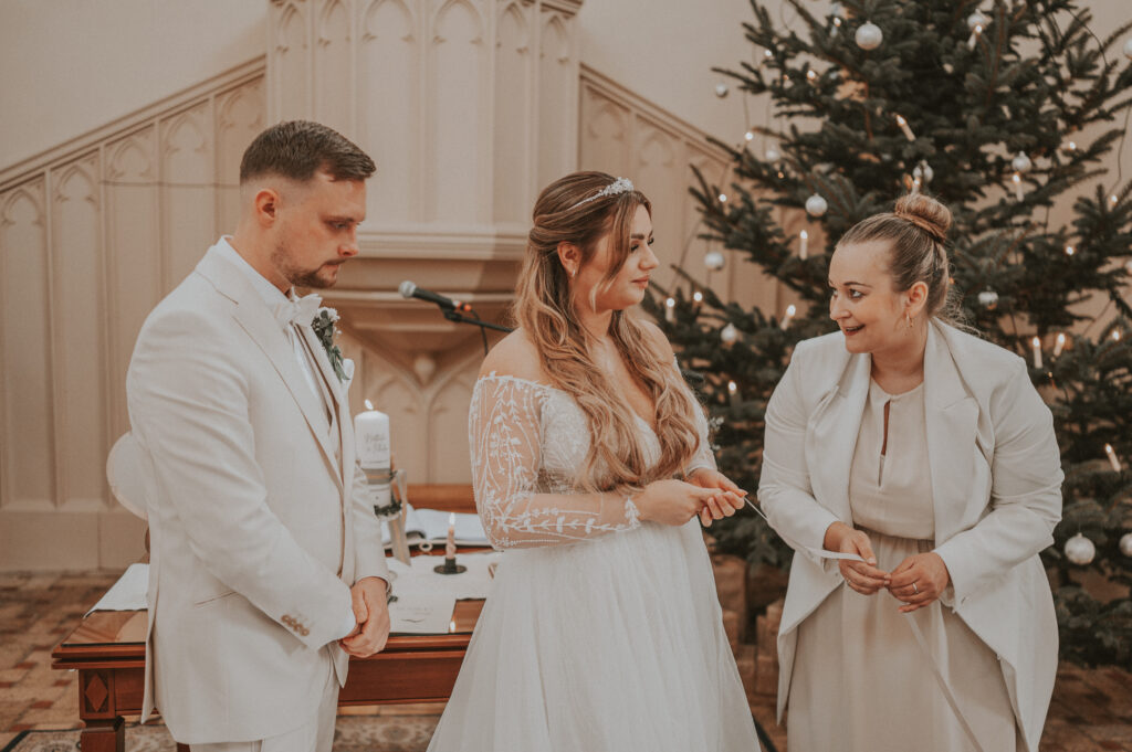 Braut und Bräutigam stehen mit ihrem Zeremonienmeister hochzeit und dem Zeremonienmeister in einer Kirche vor einem Weihnachtsbaum, alle in Weiß und Beige gekleidet.