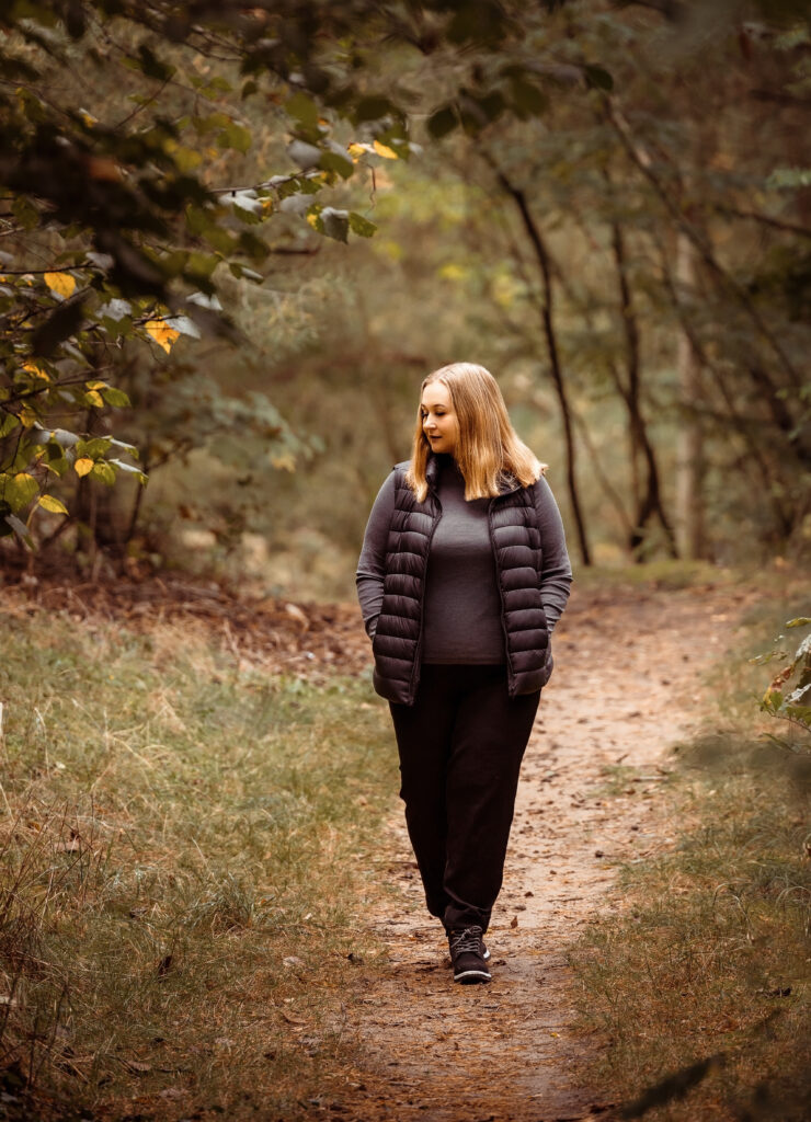 Eine Frau in einer dunklen Jacke geht allein auf einem Waldweg, umgeben von grünen Bäumen, als ob sie den perfekten Platz für eine Freie Trauung sucht.
